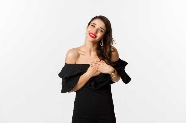 Moda y belleza. Mujer atractiva glamour en vestido negro diciendo gracias, sonriendo y cogidos de la mano en el corazón con emoción complacida, fondo blanco.