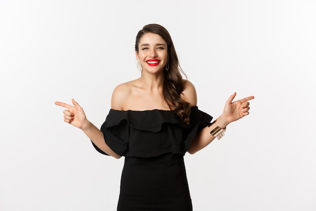 Moda y belleza. Mujer atractiva en joyería, maquillaje y vestido negro, sonriendo y señalando con el dedo hacia los lados copia oferta de espacio, fondo blanco.