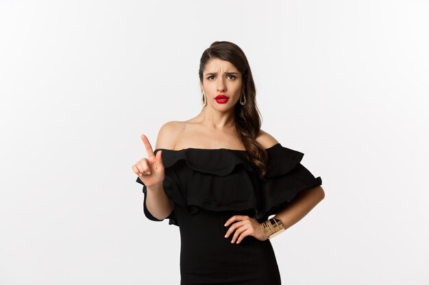 Moda y belleza. Mujer descarada en vestido negro diciendo que no, en desacuerdo y agitando el dedo disgustado, rechazando la oferta, rechazando algo, de pie sobre el fondo blanco.