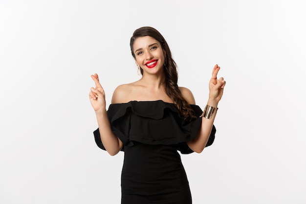 Moda y belleza. Mujer elegante glamour en vestido negro, labios rojos, luciendo optimista y sonriendo mientras cruza los dedos, pidiendo deseos, de pie sobre fondo blanco.