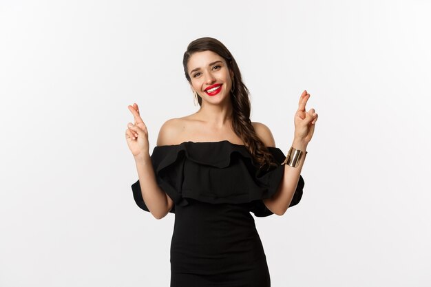 Moda y belleza. Mujer elegante glamour en vestido negro, labios rojos, luciendo optimista y sonriendo mientras cruza los dedos, pidiendo deseos, de pie sobre fondo blanco.