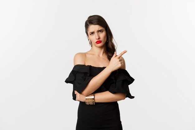 Moda y belleza. Mujer de glamour escéptica con labios rojos, vestido negro, apuntando con el dedo hacia algo aburrido y aburrido, de pie sobre fondo blanco.