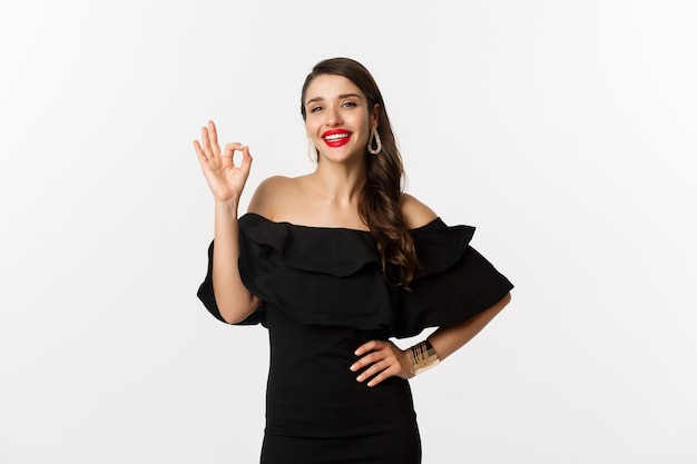 Moda y belleza. Mujer guapa satisfecha con lápiz labial rojo, vestido negro, mostrando signo bien en aprobación, como y de acuerdo, de pie sobre fondo blanco.