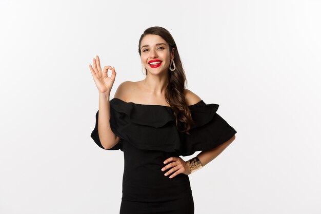 Moda y belleza. Mujer guapa satisfecha con lápiz labial rojo, vestido negro, mostrando signo bien en aprobación, como y de acuerdo, de pie sobre fondo blanco.