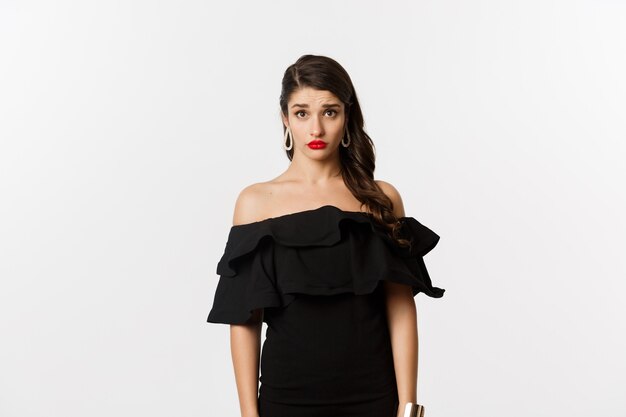 Moda y belleza. Mujer joven linda y tímida en vestido negro que parece confundida y triste a la cámara, no puedo entender, de pie triste contra el fondo blanco.