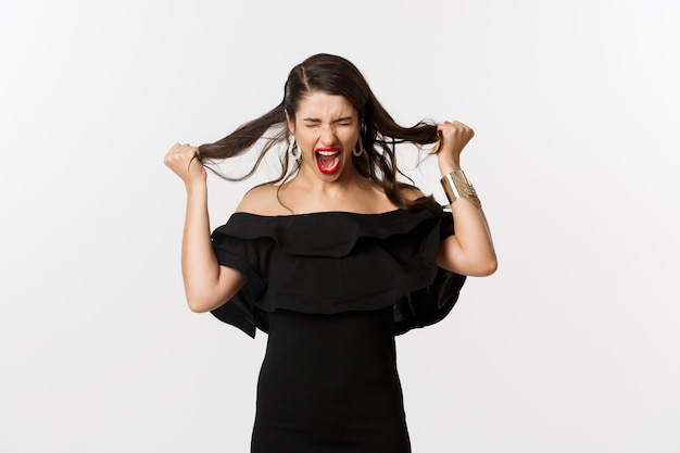 Foto gratuita moda y belleza. mujer joven en vestido negro gritando y arrancando el pelo en la cabeza, gritando enojado, de pie enojado e indignado sobre el fondo blanco.