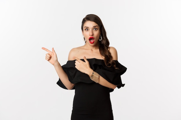 Moda y belleza. Mujer sorprendida en vestido negro glamour apuntando con el dedo hacia la izquierda, mostrando publicidad y mirando asombrado, fondo blanco