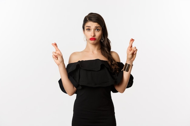 Moda y belleza. Mujer tonta esperanzada en vestido negro pidiendo deseos, sosteniendo los dedos cruzados para la buena suerte, de pie sobre fondo blanco.