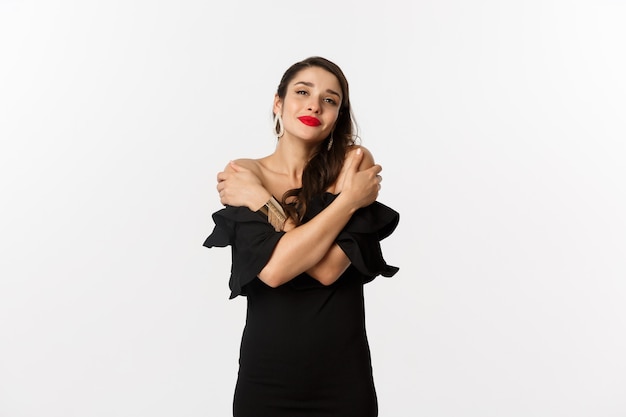 Foto gratuita moda y belleza. sensual y tierna mujer vestida de negro, abrazando su propio cuerpo, abrazándose a sí misma y sonriendo, de pie sobre fondo blanco.