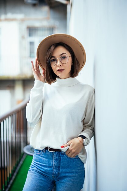 Moda callejera interesada mujer vestida con sombrero, jeans, sombrero ancho y gafas transparentes en el balcón