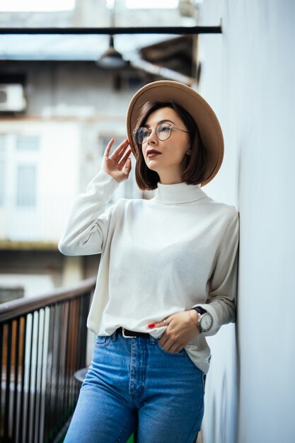 Moda callejera interesada mujer vestida con sombrero, jeans, sombrero ancho y gafas transparentes en el balcón