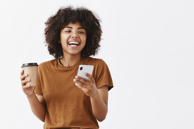 Moda despreocupada chica afroamericana con cabello rizado en camiseta marrón riendo mientras habla con amigos tomando café de una taza de papel y sosteniendo un teléfono inteligente