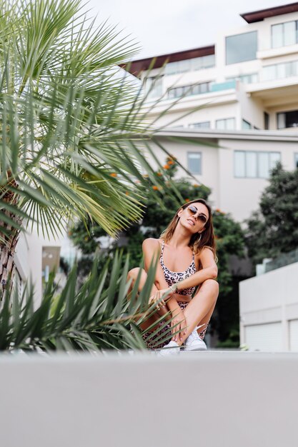 Foto gratuita moda elegante mujer europea bronceada con gafas de sol, camiseta de leopardo y pantalones cortos de motociclista, fuera de la villa cerca de la palmera