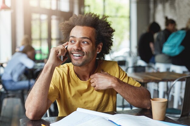 Moda hombre de piel oscura con cabello rizado con camiseta amarilla rodeado de libros que descansan en la acogedora cafetería tomando café y hablando por teléfono celular