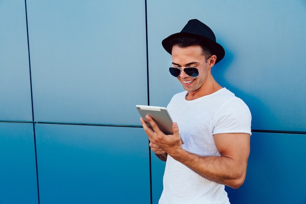 Moda joven en gafas de sol y sombrero, usando una tableta, de pie contra la pared