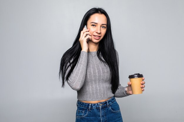 Moda joven de pie hablando por un teléfono móvil mientras sostiene una taza de café para llevar en la otra mano, aislado en la pared gris