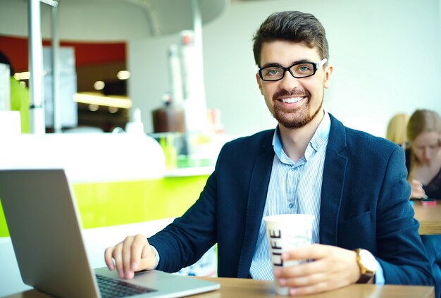 Moda joven sonriente hipster hombre tomando café en la cafetería de la ciudad durante la hora del almuerzo con cuaderno en traje