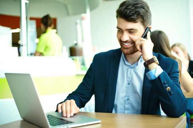 Moda joven sonriente hombre hipster en el café de la ciudad durante la hora del almuerzo con el cuaderno en traje hablando por teléfono