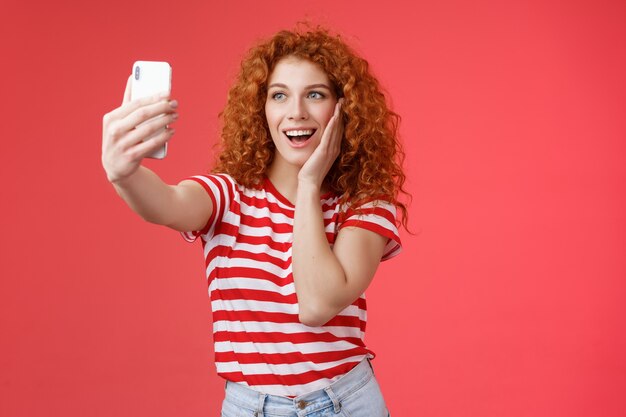 Moda linda pelirroja rizada mujer verano camiseta grabar video redes sociales de lujo tropical resolt viajes vacaciones en el extranjero tomar selfie sostener teléfono inteligente pose fotografía tonta fondo rojo.