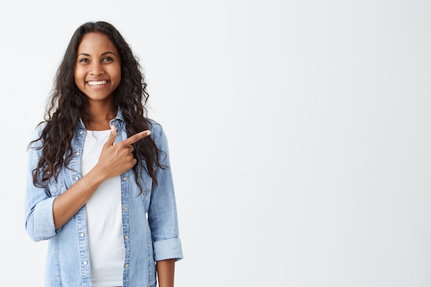 Foto gratuita moda mujer afroamericana joven emocional vistiendo camisa vaquera apuntando con su dedo índice a la pared blanca en blanco detrás de ella, mirando positiva y feliz, ampliamente sonriendo.