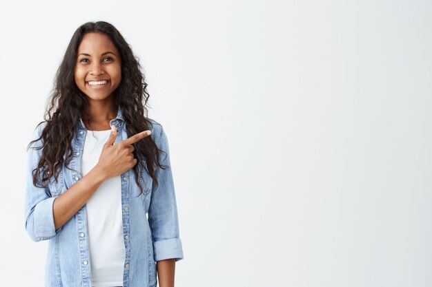 Moda mujer afroamericana joven emocional vistiendo camisa vaquera apuntando con su dedo índice a la pared blanca en blanco detrás de ella, mirando positiva y feliz, ampliamente sonriendo.