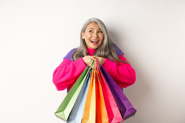 Moda mujer asiática senior adicta a las compras, abrazando bolsas de compras y sonriendo alegre, comprando con descuentos, de pie sobre fondo blanco.