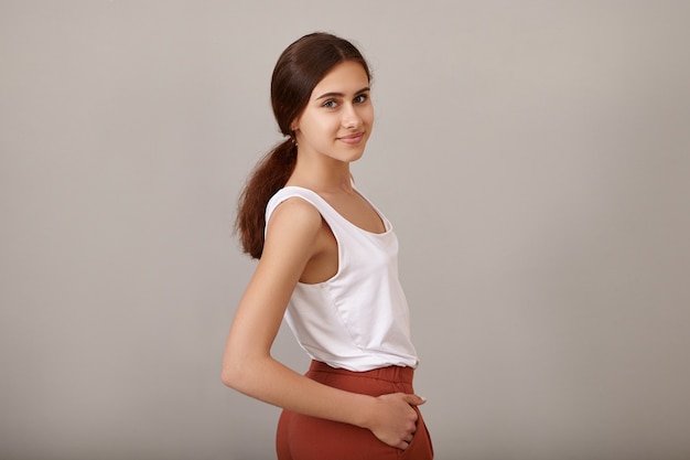 Moda mujer caucásica joven de aspecto moderno con el pelo largo y oscuro recogido en una cola de caballo desordenada posando con ropa elegante y elegante, manteniendo las manos en los bolsillos, con aspecto lúdico