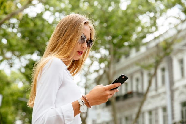 Moda mujer hermosa con largo cabello rubio usando un teléfono móvil, mientras camina al aire libre