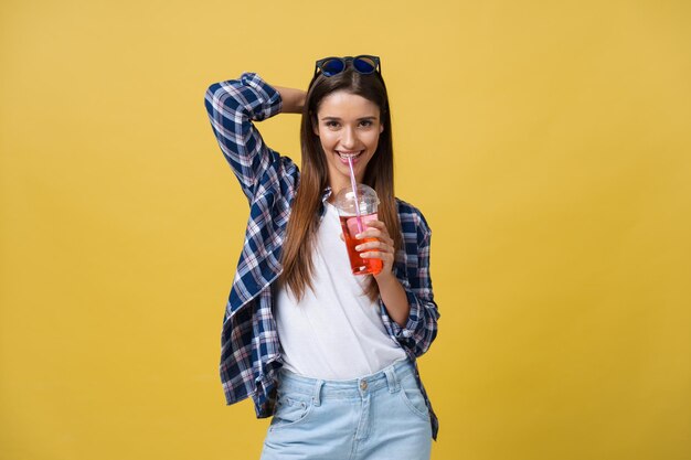 Moda mujer joven y bonita con taza de jugo de fruta fresca en camisa azul divirtiéndose sobre fondo amarillo colorido