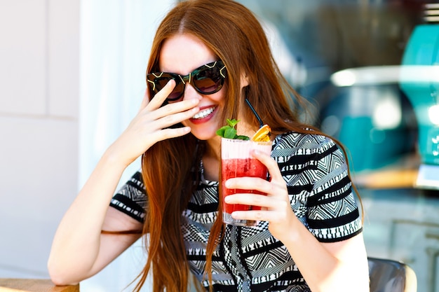 Moda mujer joven con pelos largos y una sonrisa increíble, sosteniendo una deliciosa limonada de cóctel de verano dulce, vestido elegante y maquillaje, relajándose en el café de la ciudad. felices emociones alegres.