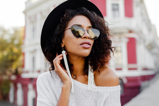 Moda mujer negra con elegantes pelos afro posando al aire libre. Fondo urbano Lleva gafas de sol negras, sombrero y aretes blancos. Accesorios de moda. Sonrisa perfecta.