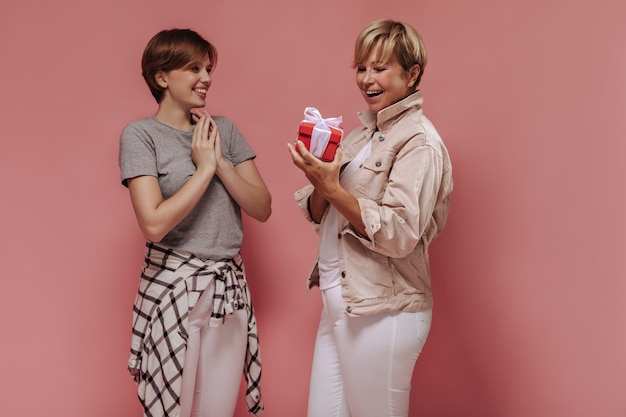 Moda mujer rubia en chaqueta beige con caja de regalo roja pequeña, sonriendo y posando con niña en camiseta gris sobre fondo rosa.