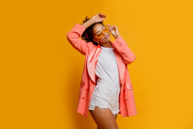Modelo africano agraciado que presenta en chaqueta de moda rosada y pantalones cortos blancos sobre fondo amarillo en estudio.