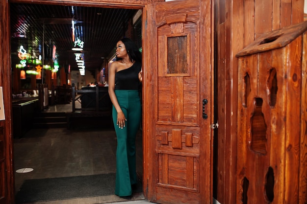 Foto gratuita modelo afroamericano delgado de belleza vestido con blusa negra y pantalones verdes de piernas largas posados en el pub