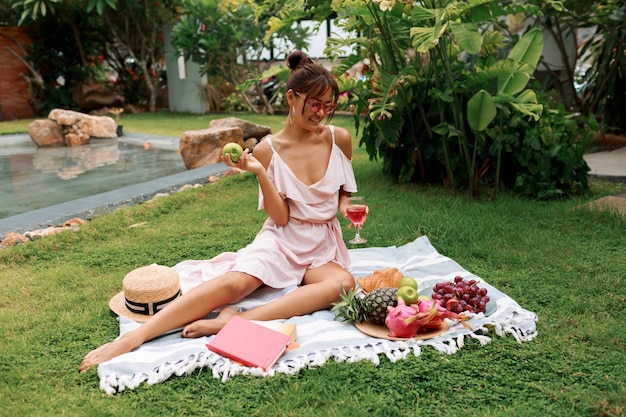 Modelo asiático agraciado sentado en una manta, bebiendo vino y disfrutando de un picnic de verano en un jardín tropical.