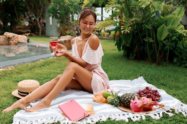 Modelo asiático bastante elegante sentado en una manta, bebiendo vino y disfrutando de un picnic de verano en un jardín tropical.