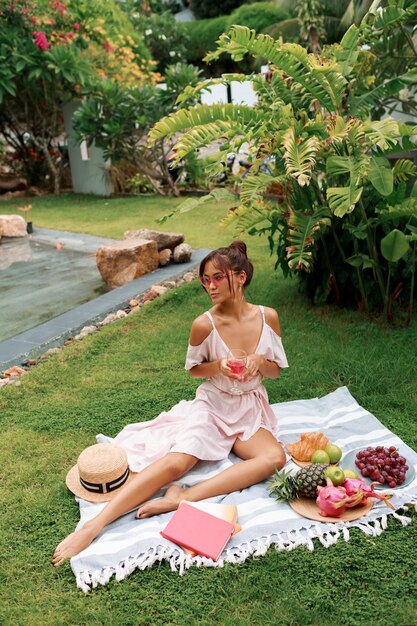 Modelo asiático elegante y romántico sentado en una manta, bebiendo vino y disfrutando de un picnic de verano en un jardín tropical.