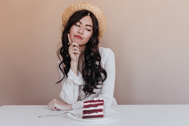 Modelo asiático pensativo mirando la torta. Dudando a la mujer japonesa posando con postre.