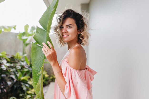 Modelo bastante femenino con ojos azules mirando y sosteniendo la hoja verde. Adorable chica bronceada con expresión de cara tranquila de pie en el patio.