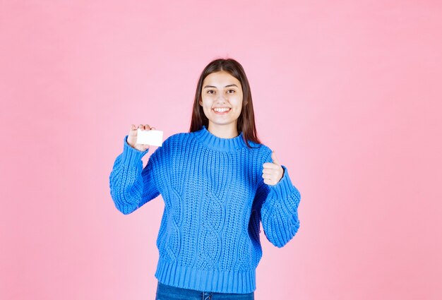 modelo de chica joven con una tarjeta que muestra un pulgar hacia arriba en la pared rosa.