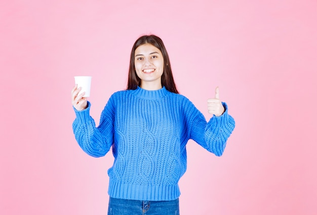 modelo de chica joven con un vaso de plástico mostrando un pulgar hacia arriba en la pared rosa.