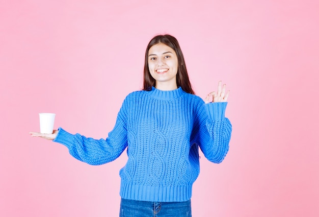 modelo de chica joven con un vaso de plástico que muestra un gesto bien en la pared rosa.