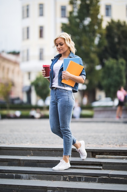 Modelo de chica rubia va a las clases de trabajo a través del centro de la ciudad sosteniendo una computadora portátil de café en sus manos por la mañana