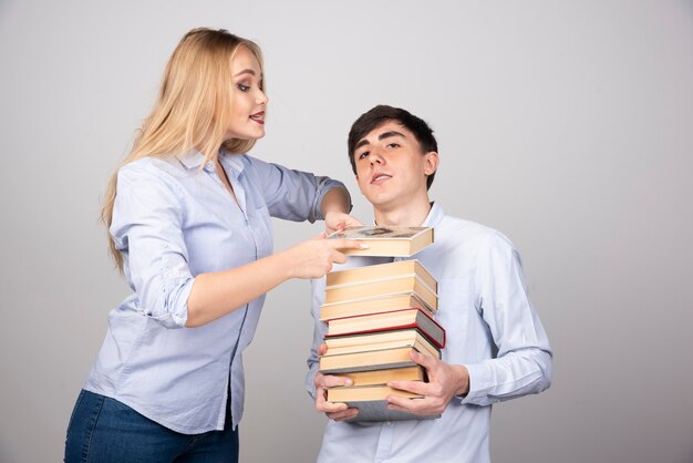 Modelo de chico morena de pie y llevando una pila de libros cerca de mujer rubia