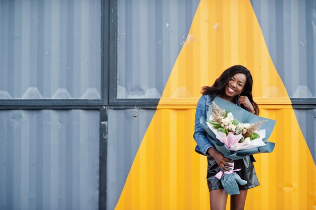 Modelo delgada afroamericana de piel oscura posada en pantalones cortos de cuero negro y chaqueta de jeans Sostiene un ramo de flores contra una pared de acero triangular amarilla