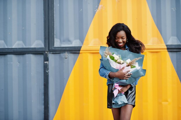 Modelo delgada afroamericana de piel oscura posada en pantalones cortos de cuero negro y chaqueta de jeans Sostiene un ramo de flores contra una pared de acero triangular amarilla