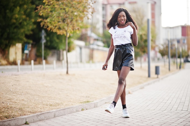 Modelo delgado afroamericano de piel oscura posado en pantalones cortos de cuero negro y camiseta blanca