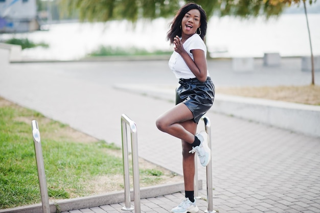 Modelo delgado afroamericano de piel oscura posado en pantalones cortos de cuero negro y camiseta blanca