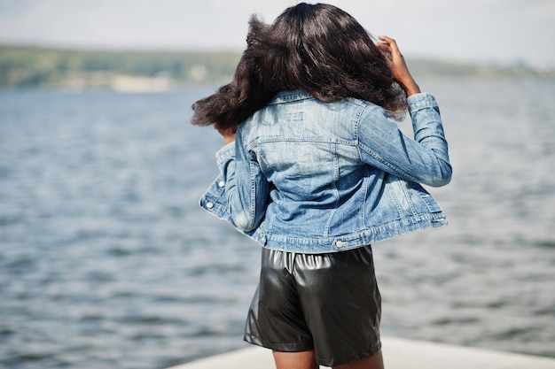 Modelo delgado afroamericano de piel oscura posado en pantalones cortos negros y chaqueta de jeans contra el mar