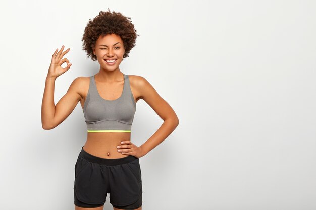 Modelo deportivo en ropa deportiva que muestra un buen gesto con la mano, asegura que todo está bien, mantiene la otra mano en la cintura